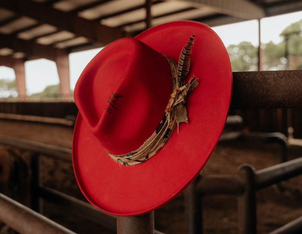 A red cowboy hat handing on a fence inside a barn.